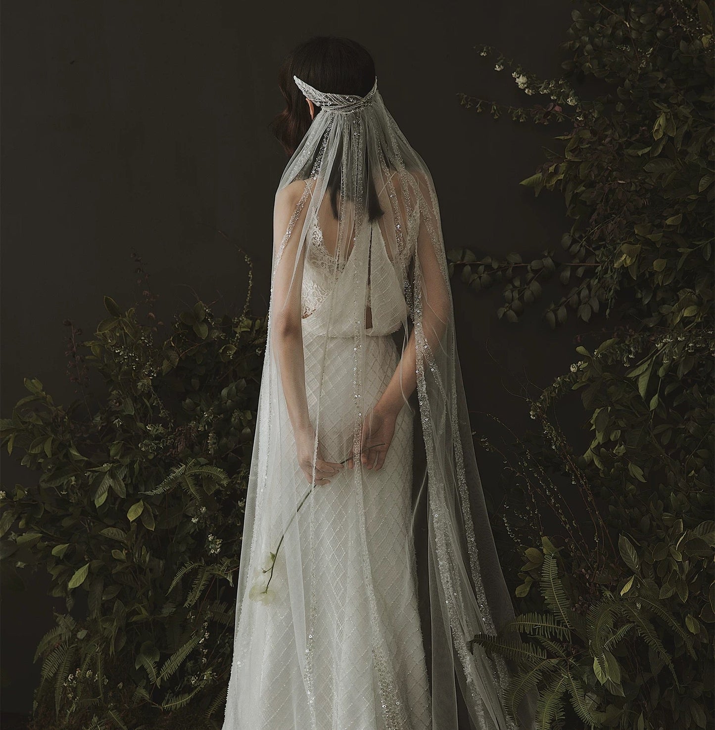 Person wearing a white wedding dress with a veil in a dark, leafy setting, featuring the brand 'DearWhite'.