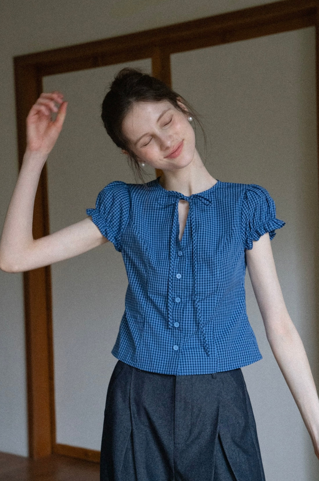Woman wearing a blue checkered blouse with puffed sleeves in a minimal indoor setting.