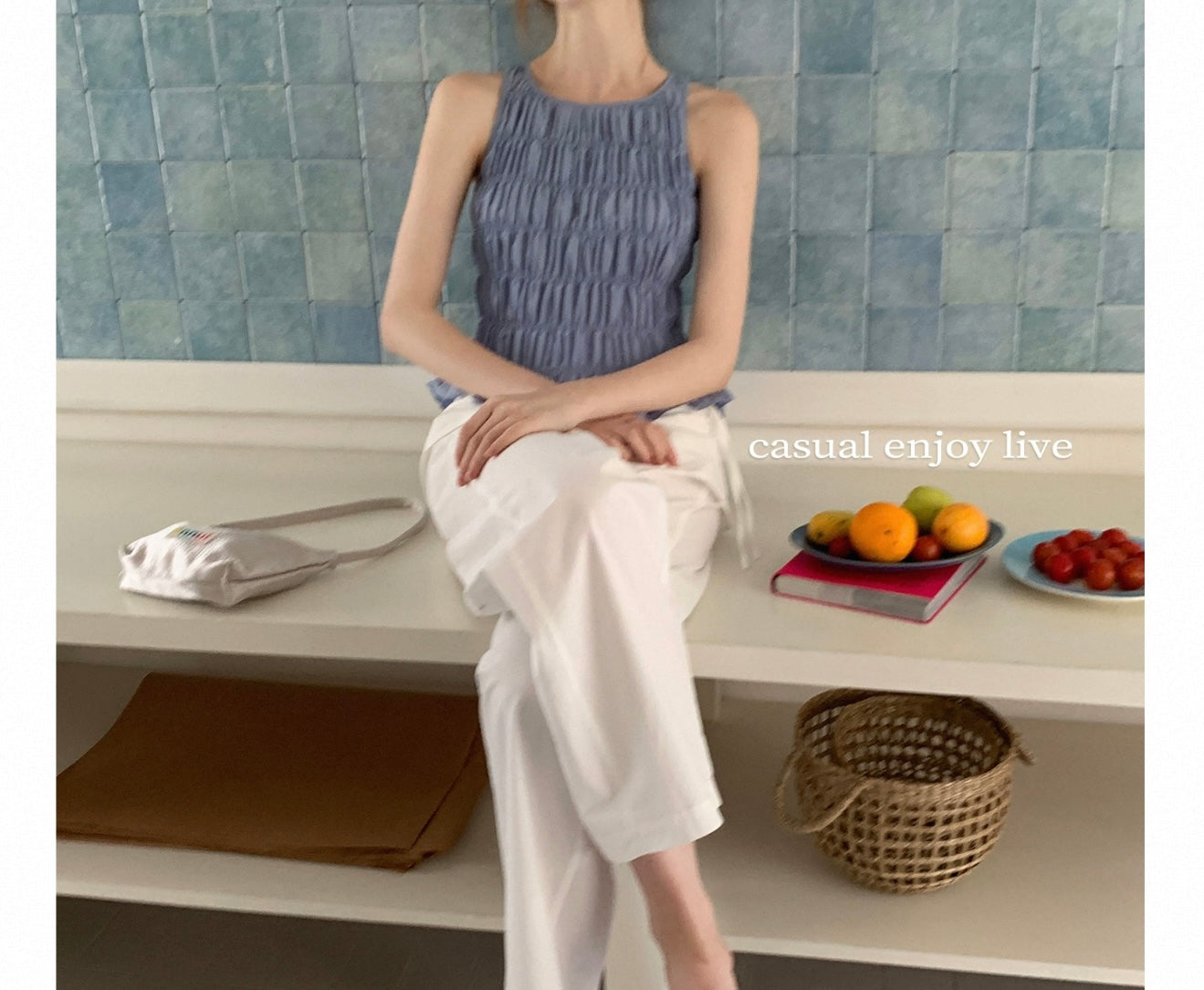 Person sitting on a bench with a casual pose, wearing a blue top and white pants, in front of a tiled wall.