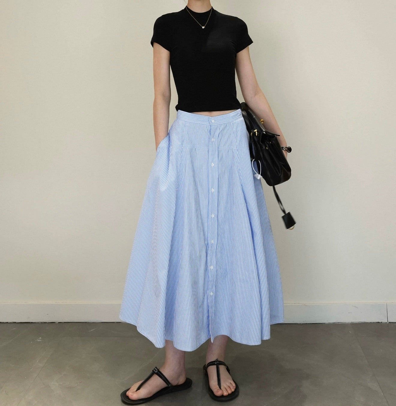 Person wearing a black top and blue striped skirt holding a black bag against a plain background