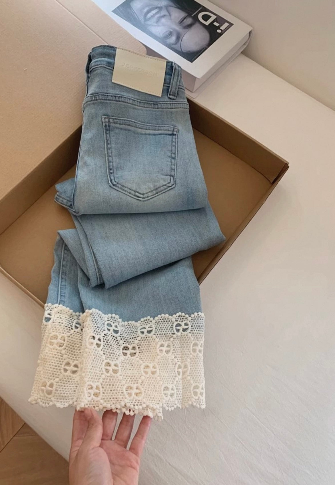 Folded light blue jeans with lace trim held above a cardboard box.