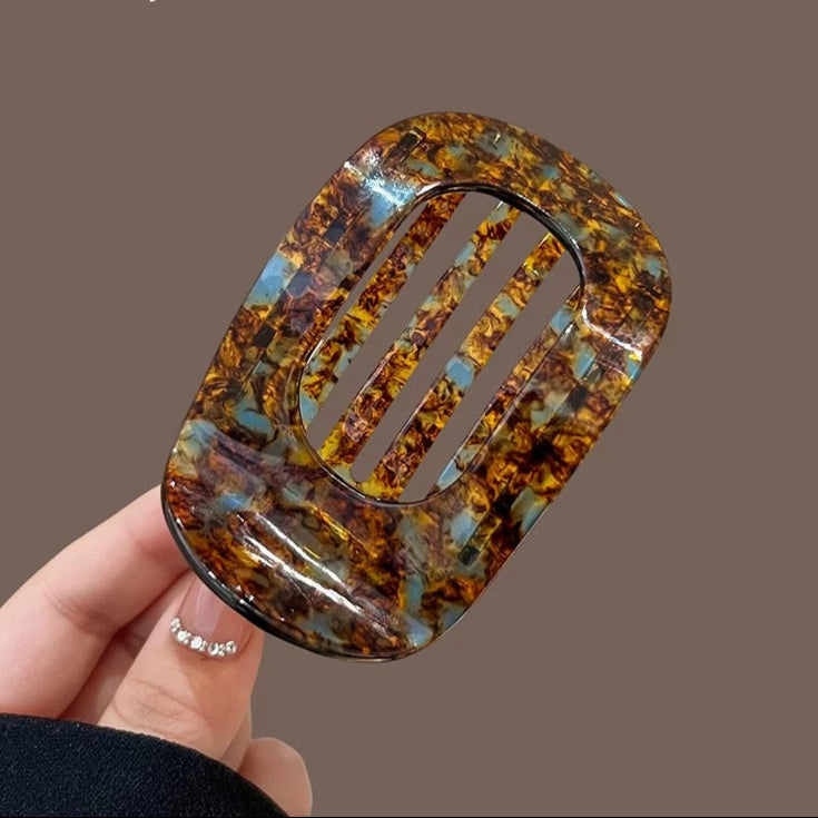 Hand holding a decorative hair clip with a brown and blue pattern against a brown background