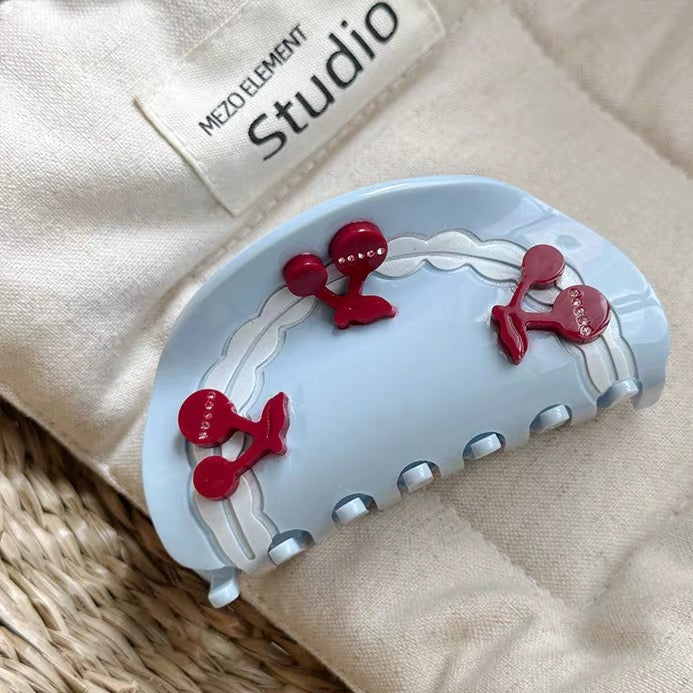 Blue hair clip with red details on a beige fabric background, featuring branding.
