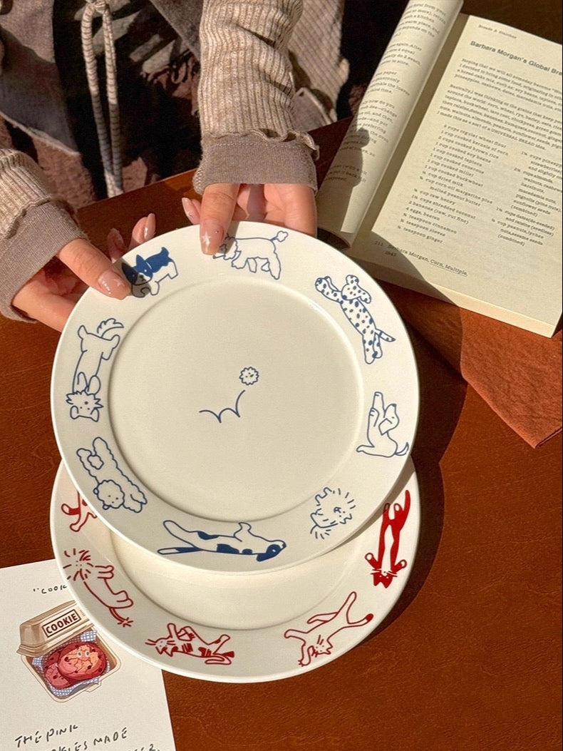 Two ceramic plates with animal illustrations held by a person on a wooden table, with a book and card in the background.