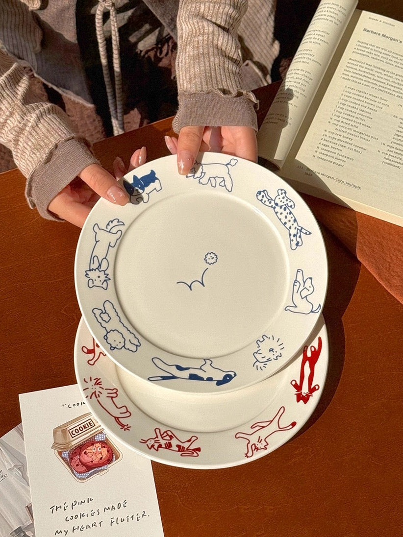 Two ceramic plates with animal illustrations held by a person on a wooden table, with a book and card in the background.