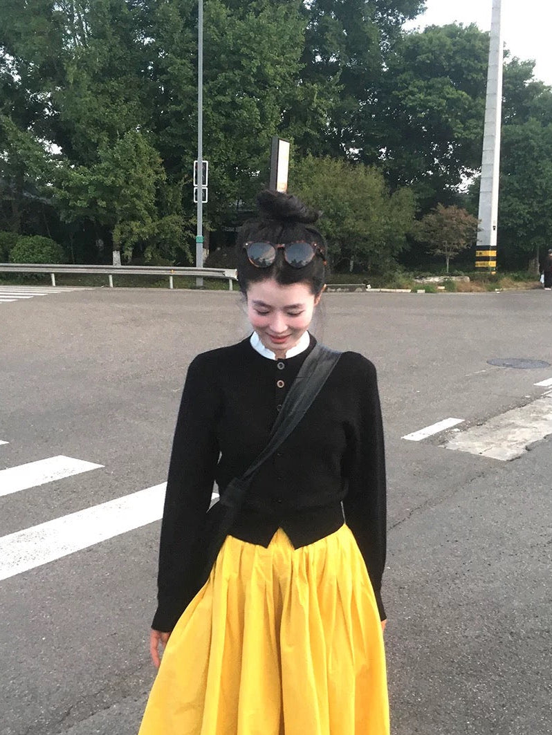 Woman in black top and yellow skirt standing on a road with trees in the background, wearing sunglasses on her head.