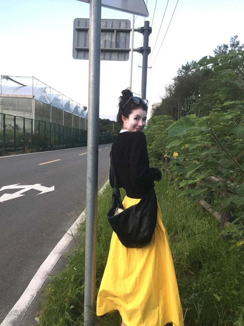 Person wearing a black top and yellow skirt standing next to a road with greenery in the background, with 'VINONITA' branding.