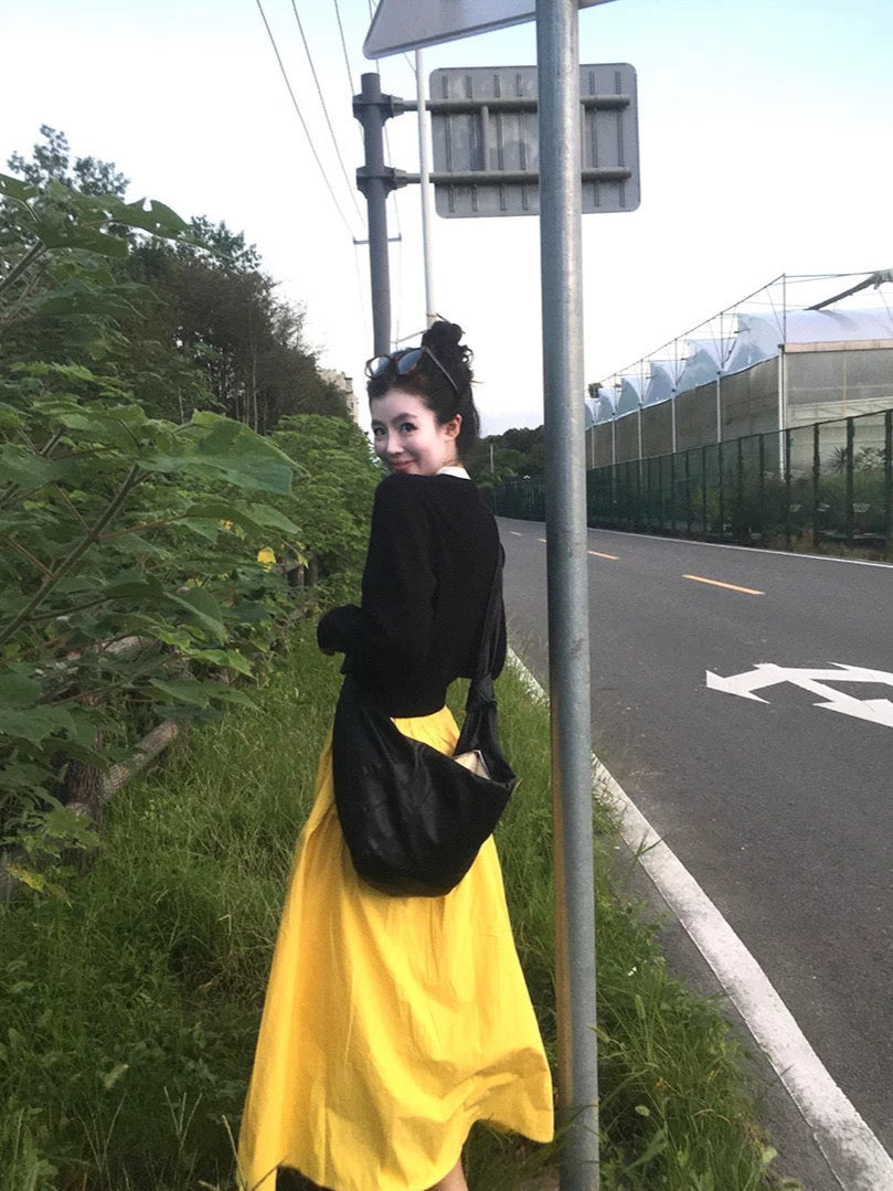 Woman in black top and yellow skirt standing on a roadside with greenery and road signs in the background.