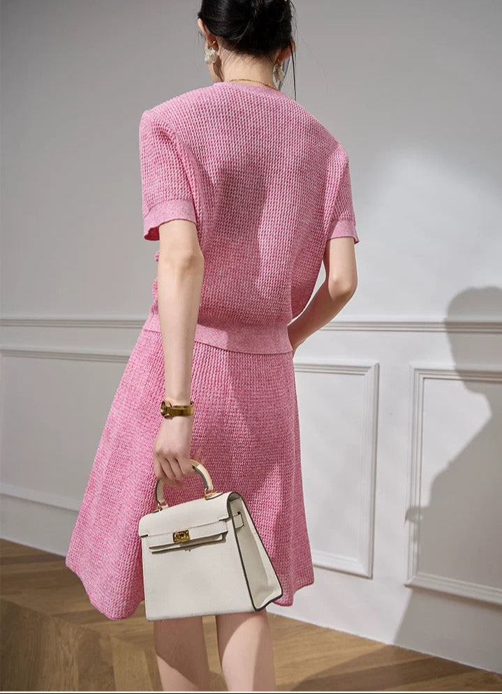 Woman in a pink dress holding a white handbag against a neutral wall.