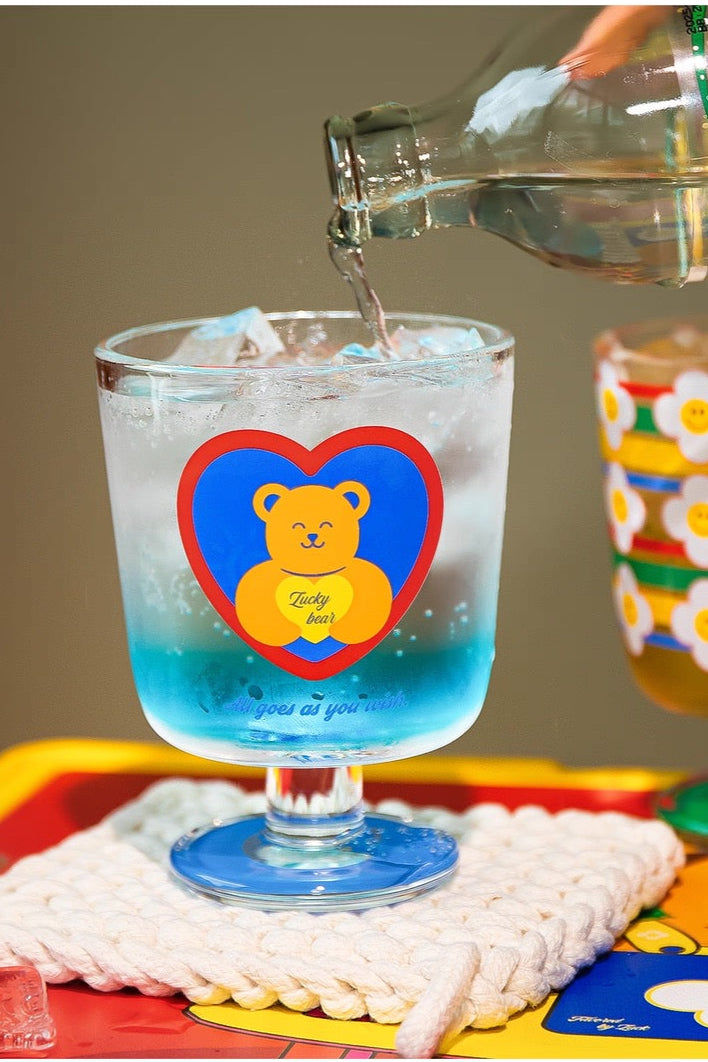 Glass with a teddy bear design being filled with a clear liquid on a colorful tray.