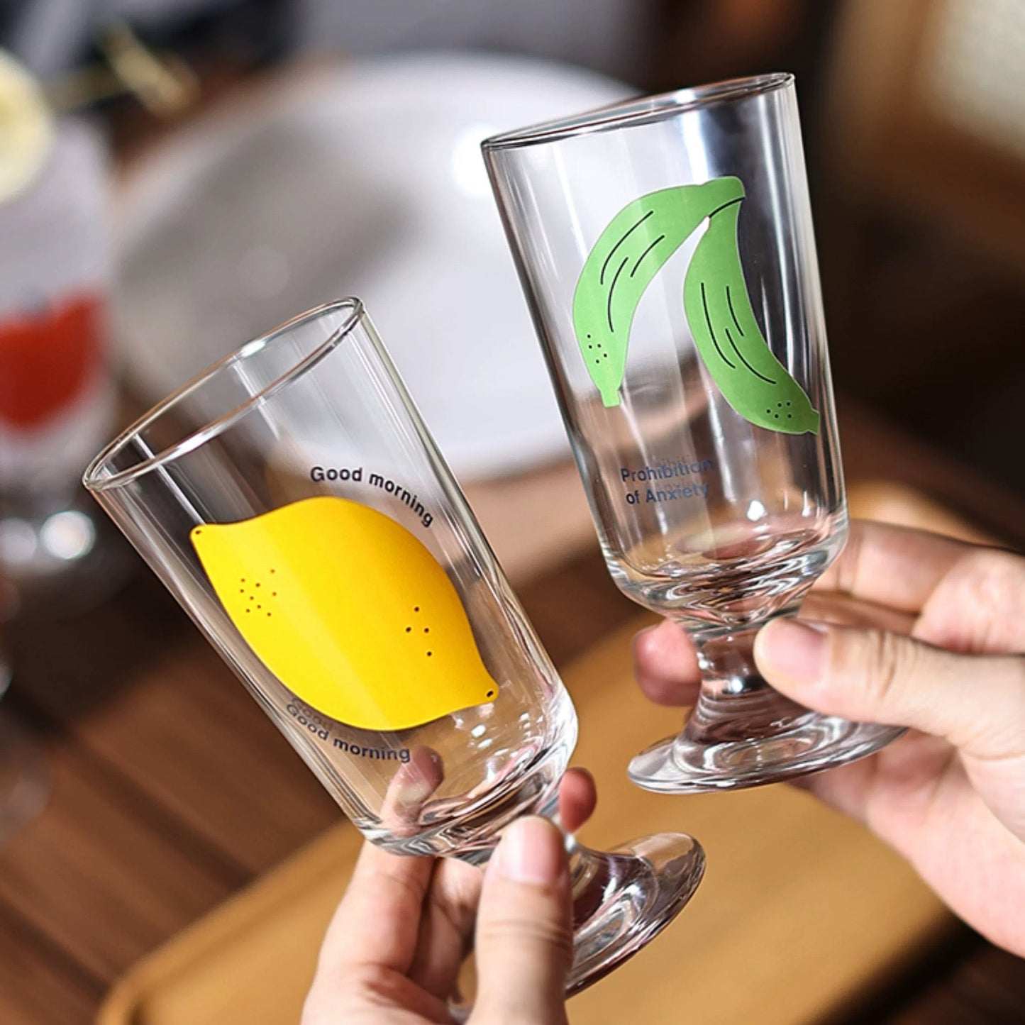 Two clear glasses with lemon and banana designs held by hands on a blurred background.