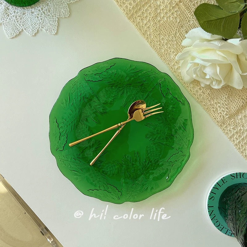 Green decorative plate with gold spoon on a white surface, surrounded by greenery and flowers.