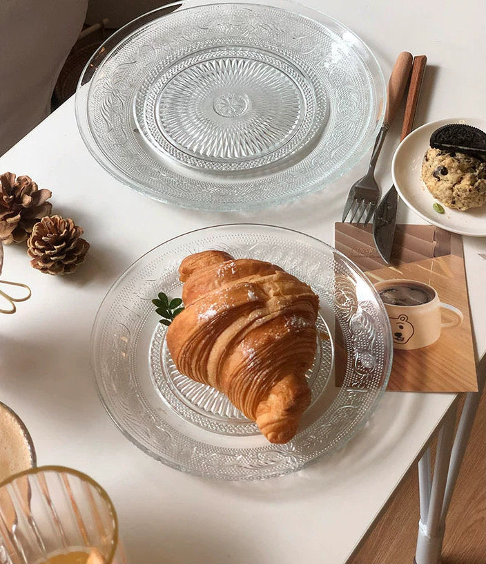 Glass plate with a croissant on a white table with a coffee cup and cookies.