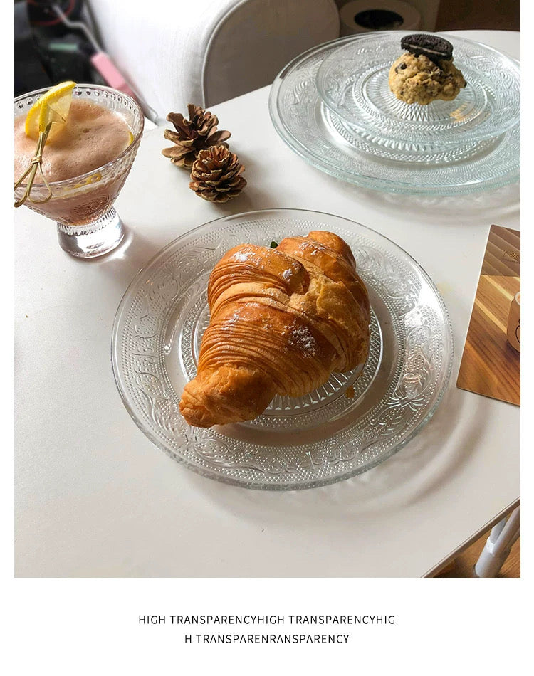 Croissant on a transparent glass plate with a drink and decorative elements on a white surface.