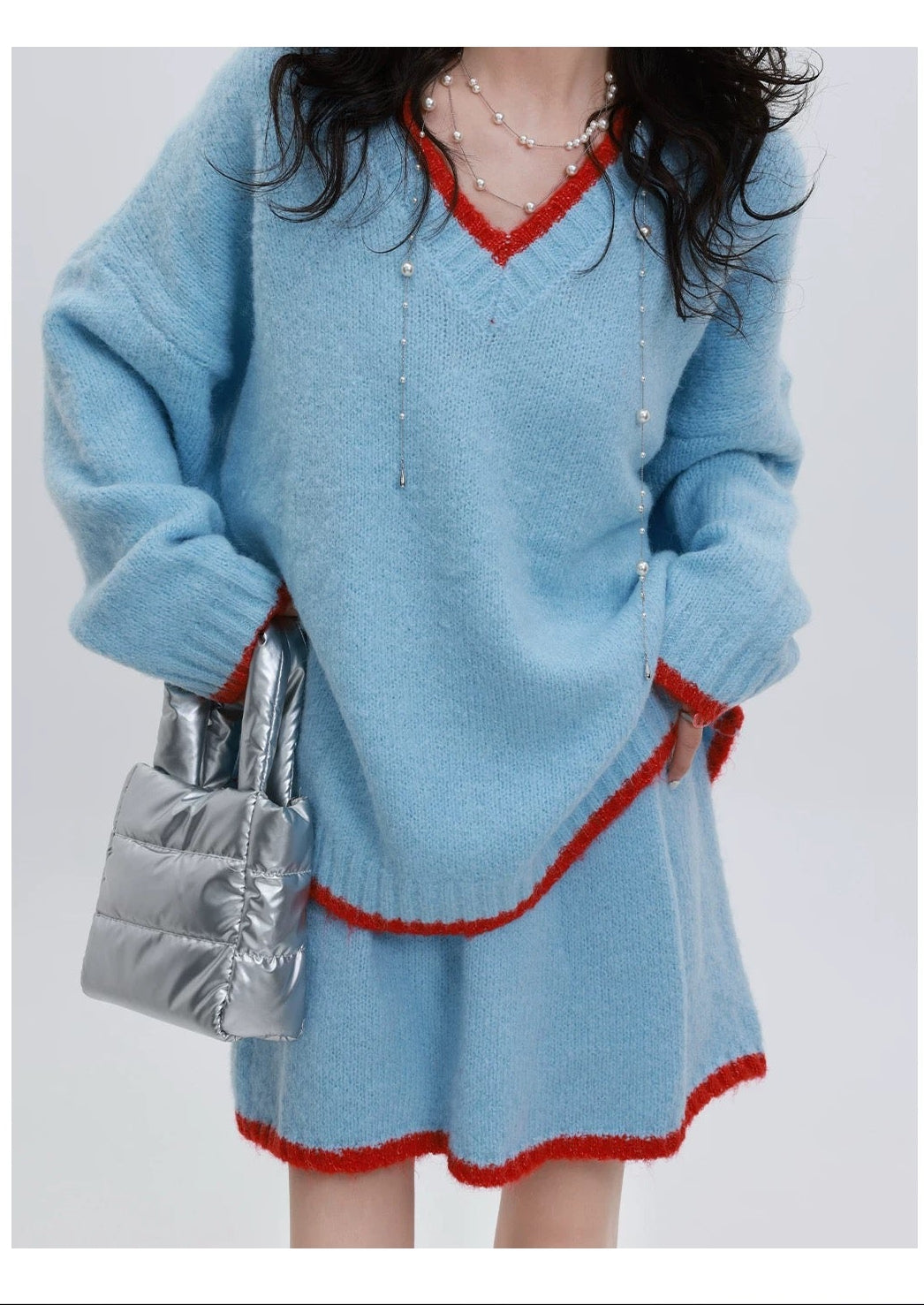 Person wearing a light blue sweater with red trim and holding a silver bag on a white background