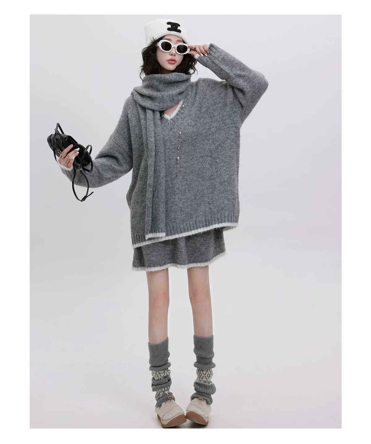 Person wearing a gray oversized sweater with a scarf, holding sunglasses, on a white background