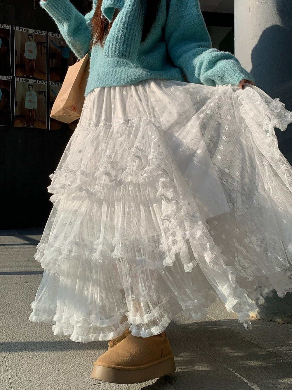 Person wearing a turquoise sweater and white lace skirt with brown boots, standing on a street.