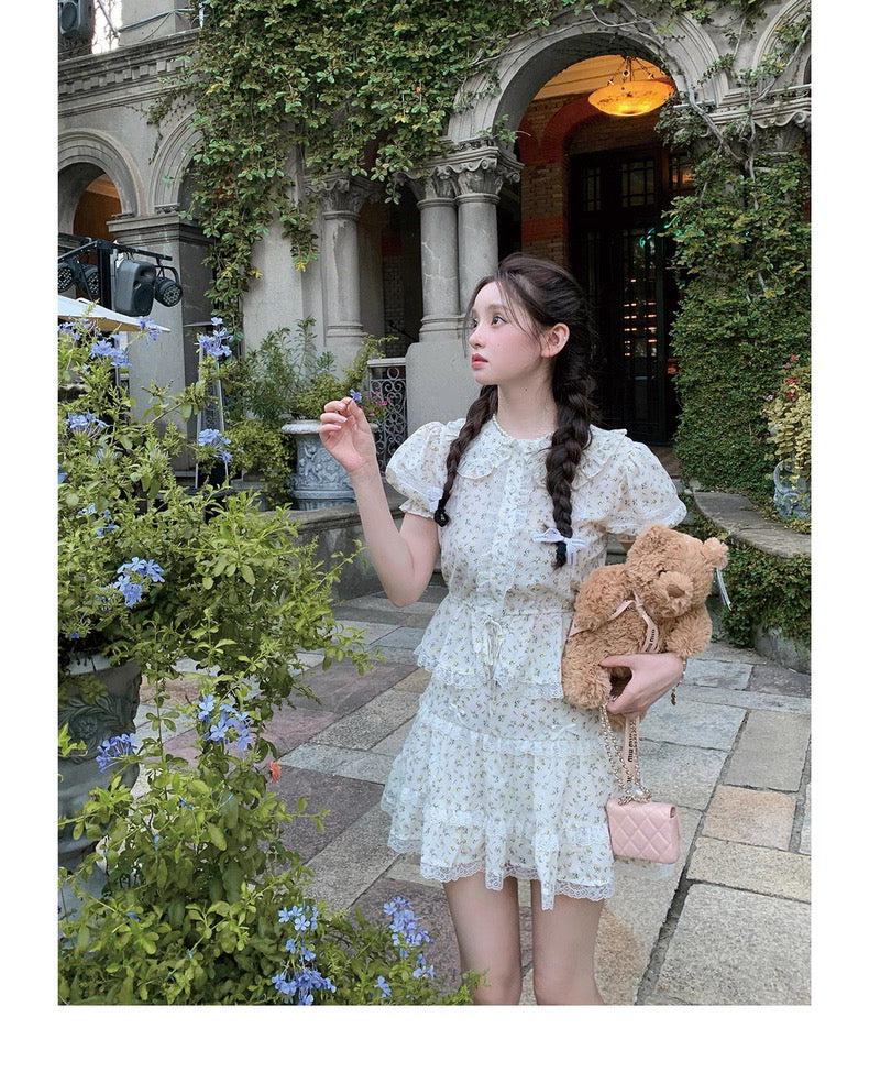 Woman in a white lace dress holding a teddy bear and pink handbag in an elegant outdoor setting.