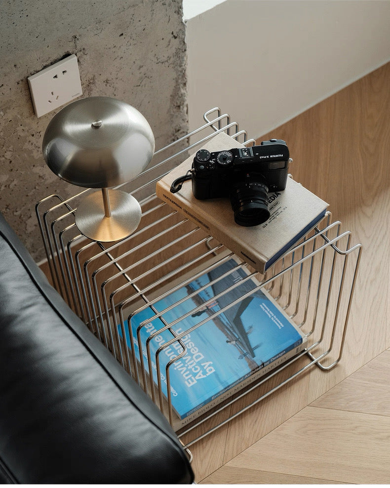 Wire magazine rack with books, a camera, and a lamp on a wooden floor.