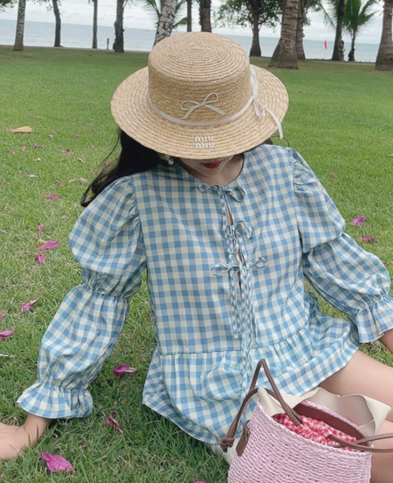 Person wearing a straw hat and blue checkered dress sitting on grass with palm trees in the background