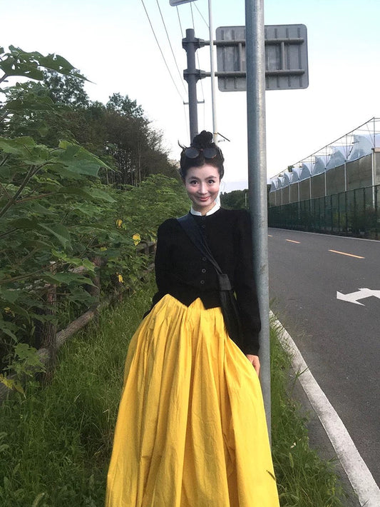 Person wearing a black top and yellow skirt standing on the side of a road with greenery in the background.