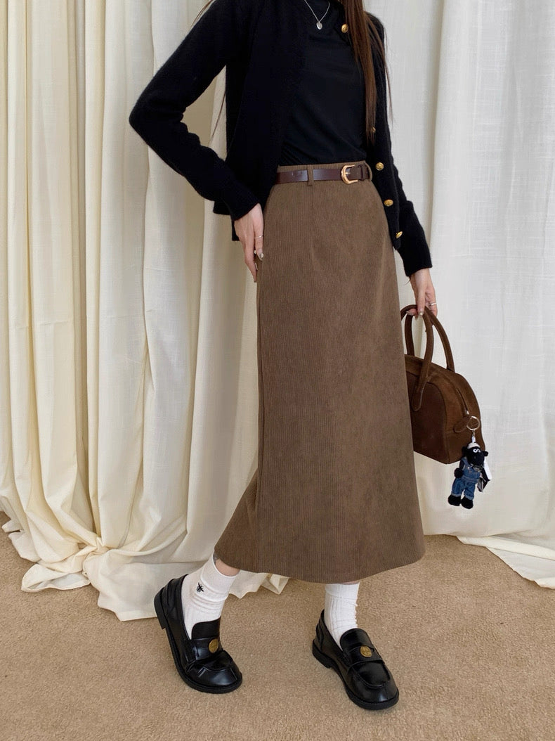 Person wearing a black top and brown skirt with a handbag against a white curtain background